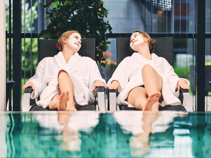 To kvinder i hvide gevandter med ansigtsmasker slapper af i liggestole ved en indendørs pool og smiler til hinanden.