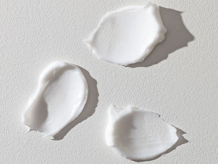 Tre udstrygninger af hvid creme eller lotion på en struktureret lysegrå overflade, set ovenfra.
