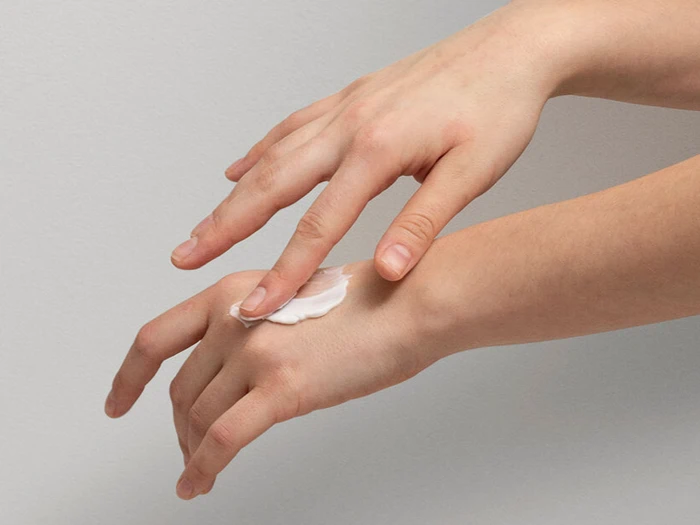 En person smører en klat hvid lotion på håndryggen ved hjælp af fingrene på den anden hånd mod en almindelig baggrund.