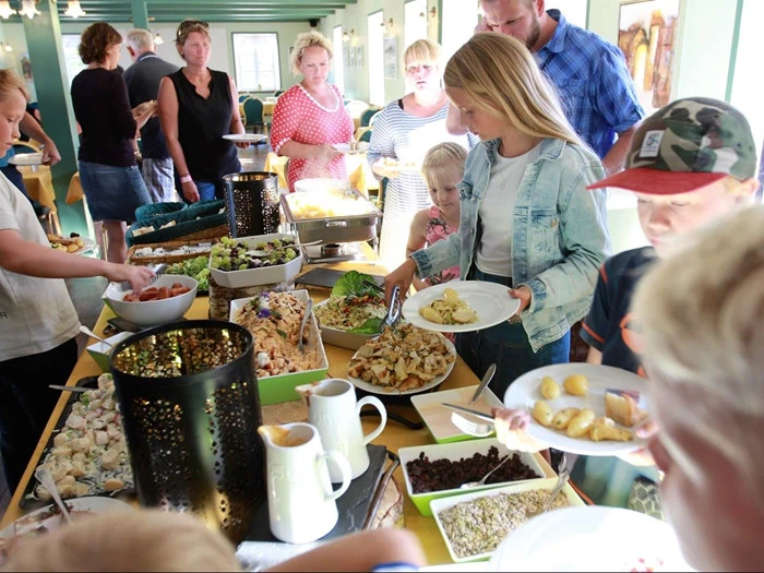 En gruppe mennesker serverer mad for sig selv fra et buffetbord fyldt med forskellige retter i et oplyst spiseområde.