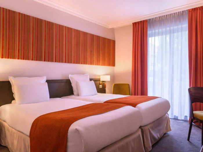 Et hotelværelse med to enkeltsenge, orange plaider, hvidt sengetøj, en stribet vægdekoration, et vindue med gennemsigtige og orange gardiner og en lille lampe på natbordet.