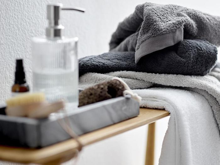 På en træbænk ligger sirligt foldede grå håndklæder og en bakke med en sæbedispenser i glas, en lille flaske, en børste og en svamp.