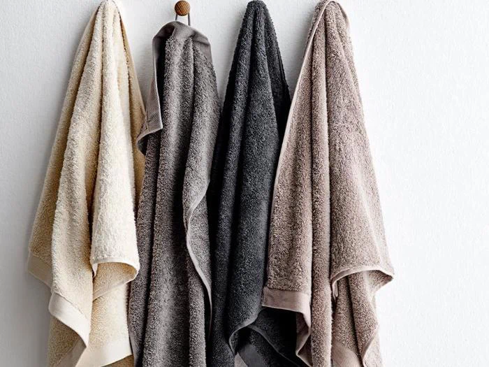 Fire håndklæder i beige, grå, mørkegrå og taupe hænger side om side på kroge mod en hvid væg.