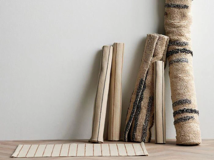 Fem sammenrullede neutralfarvede tæpper med forskellige enkle mønstre læner sig op ad en hvid væg; et udrullet stribet tæppe ligger fladt på et lyst trægulv.