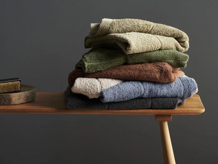 En pænt foldet stak håndklæder i forskellige farver - beige, grøn, brun og blå - ligger på en træbænk mod en grå baggrund.