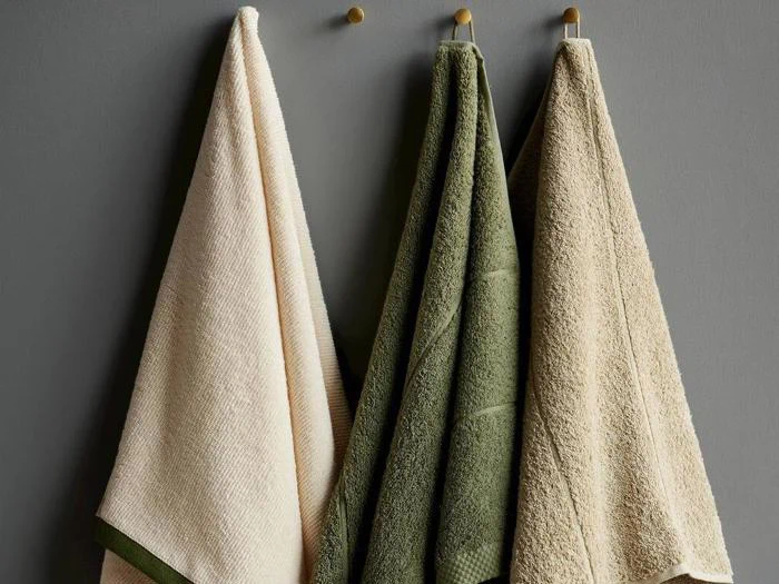 Tre håndklæder - to beige og et grønt - hænger på kroge op ad en grå væg. Hvert håndklæde er struktureret og let draperet.