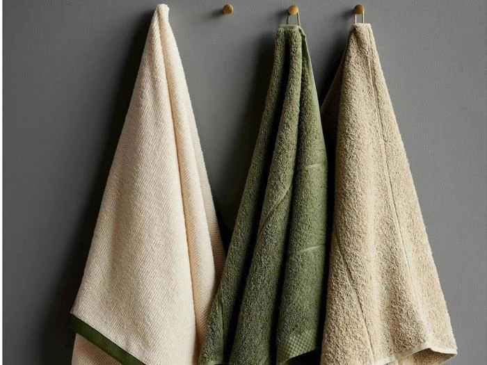 Tre håndklæder - et cremefarvet, et grønt og et beige - hænger på kroge op ad en grå væg.