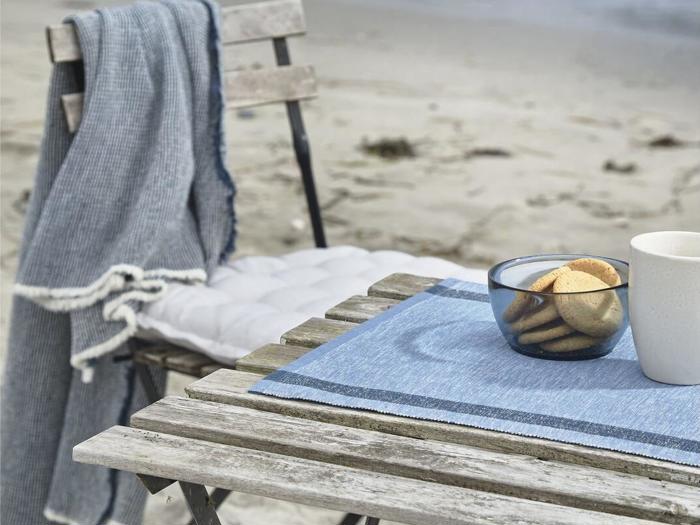 Et rustikt træbord og en stol på en strand med et blåt klæde, småkager i en skål, et krus og et blåt tæppe over stolen.