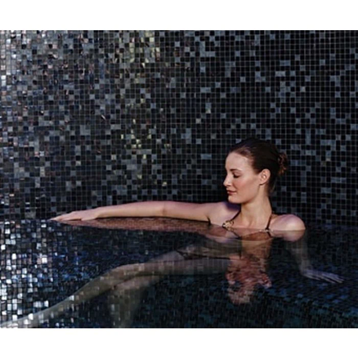 En kvinde i sort badedragt slapper af i en pool med mørke mosaikfliser, hendes arm hviler på poolkanten, og hendes blik er nedadvendt.