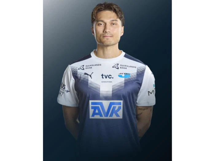 En mandlig fodboldspiller i en blå og hvid trøje står foran en mørk baggrund med farveovergang og ser ind i kameraet med hænderne bag ryggen.