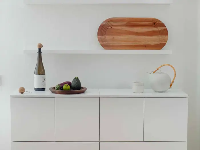 Et minimalistisk hvidt skab med en vinflaske, en træskål med frugt, en hvid tekande og en kop på toppen; træbakke på en hylde ovenover.