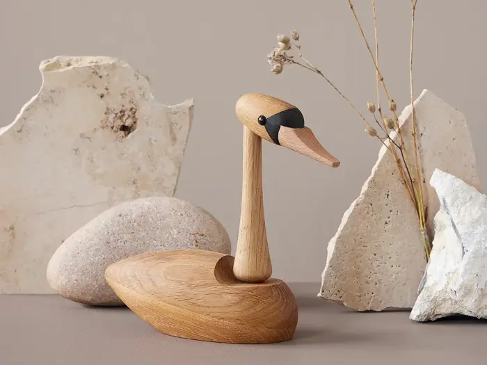 En fuglefigur af træ med lang hals sidder på en flade, omgivet af lyse sten og tørrede grene på en beige baggrund.