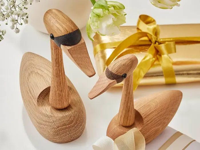 To andefigurer af træ med sorte aftegninger står ved siden af en guldindpakket gave og hvide blomster på en lys overflade.