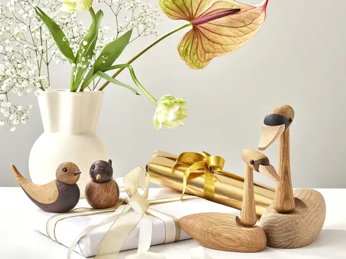 Fugle- og svanefigurer af træ sidder ved siden af hvide og guldindpakkede gaver og en hvid vase med blomster på en lys overflade.