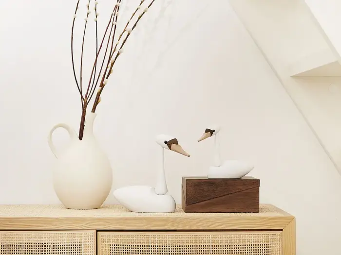 En hvid keramisk kande med høje grene og to svanefigurer i træ står på et flettet skab mod en hvid væg.