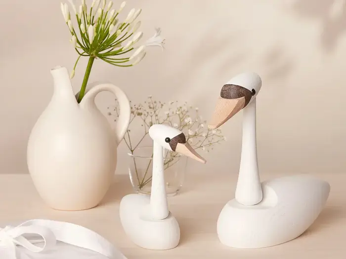 To svanefigurer af træ står på en lys flade ved siden af en hvid keramikkande med grønne blomster og en glasvase med små hvide blomster.
