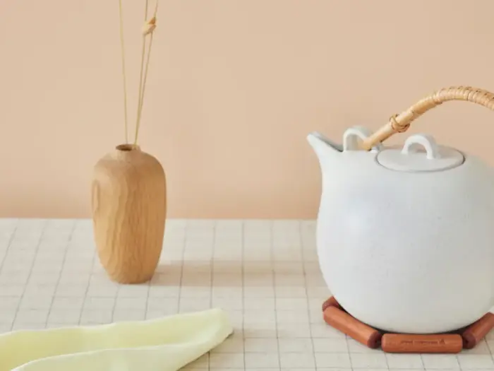 En hvid tekande med bambushåndtag står på et bord ved siden af en lille trævase med sivdiffusorer og en foldet lysdug.