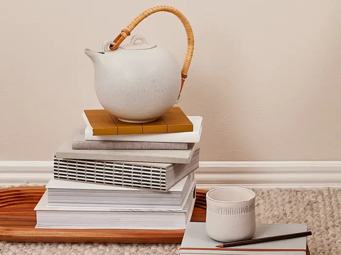 En keramisk tekande med en flettet hank står på stablede bøger ved siden af en keramisk kop og en blyant, alt sammen på en træbakke og et gulvtæppe.