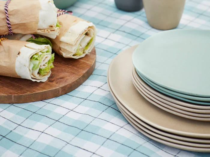 Pak sandwich ind på et træbræt ved siden af stabler af beige og lyseblå tallerkener på en blå og hvid ternet dug.