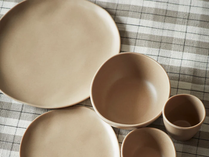 Neutralfarvede keramiktallerkener, skåle og en kop arrangeret på en beige og grå ternet dug.