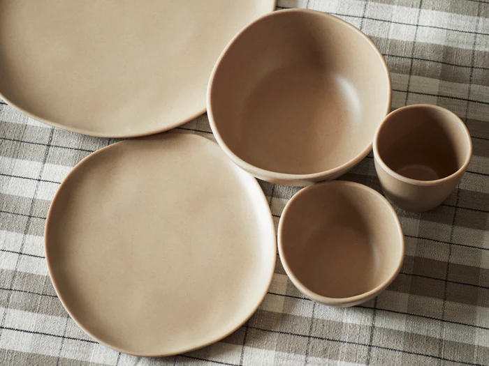 Beige keramiktallerkener, skåle og kopper arrangeret på en ternet dug i neutrale toner.