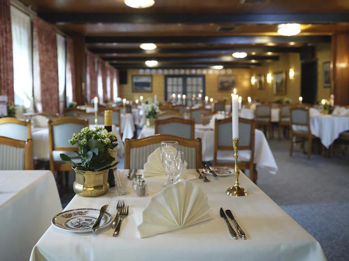 Elegant restaurantspisestue med hvide duge, foldede servietter, stearinlys, glas og potteplanter på bordene, dæmpet belysning og rækker af stole.