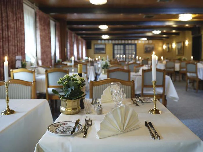 Elegant restaurantspisestue med borde til to personer med hvide duge, foldede servietter, stearinlys, blomster og sirligt arrangeret bestik og glas.