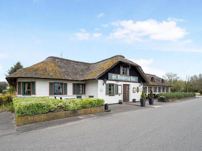 En hvid stråtækt bygning med grønne kanter og et skilt med teksten St. Aidenvej Kro står ved siden af en vej under en blå himmel med grønne områder foran.
