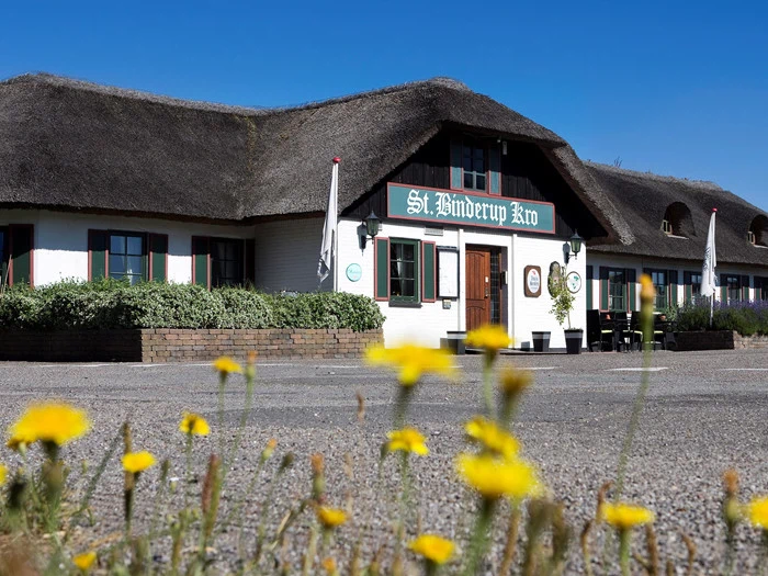 En bygning med stråtag og et skilt med teksten Skt. Binderup Kro, omgivet af grønne områder og gule vilde blomster i forgrunden under en klar blå himmel.