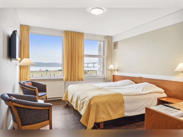 Et hyggeligt hotelværelse med en dobbeltseng, to stole, et lille bord, tv på væggen og et stort vindue med gule gardiner med udsigt over et naturskønt område.