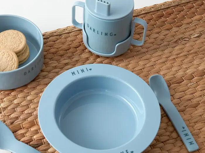 Blåt børnefad med MINI- og DARLING-tekst, bestående af skål, kop med hank, tallerken, gaffel og ske på en vævet måtte; småkager i en skål.