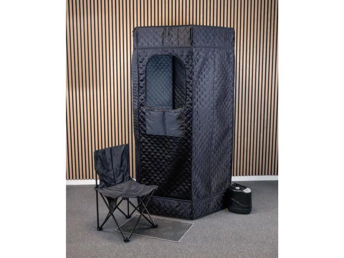 Sort quiltet privattelt med vindue, sammenklappelig sort stol og et lille sort apparat på gulvet mod en stribet trævæg og et gråt gulvtæppe.