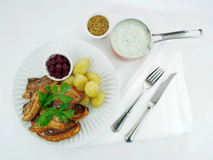 En tallerken med sprød flæskesvær, kogte kartofler, rødbeder i tern, pyntet med persille. Ved siden af står en kop sennep, cremet sauce i en gryde, gaffel og kniv på en hvid serviet.