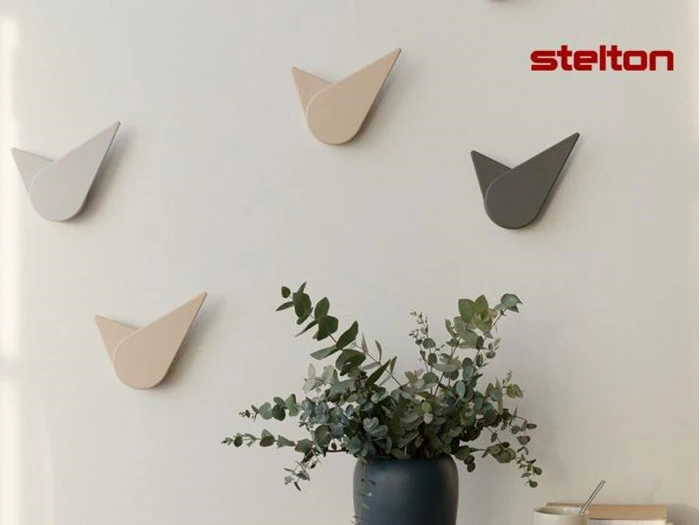 Minimalistiske vægure i neutrale toner er arrangeret på en hvid væg over en grøn vase med eukalyptusgrene; stelton-logoet er i øverste højre hjørne.
