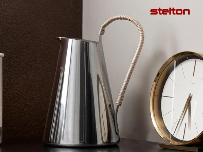 En skinnende kande i rustfrit stål med et buet, indpakket håndtag står ved siden af et rundt ur i guld og hvidt. Ordet stelton står med rødt i øverste højre hjørne.