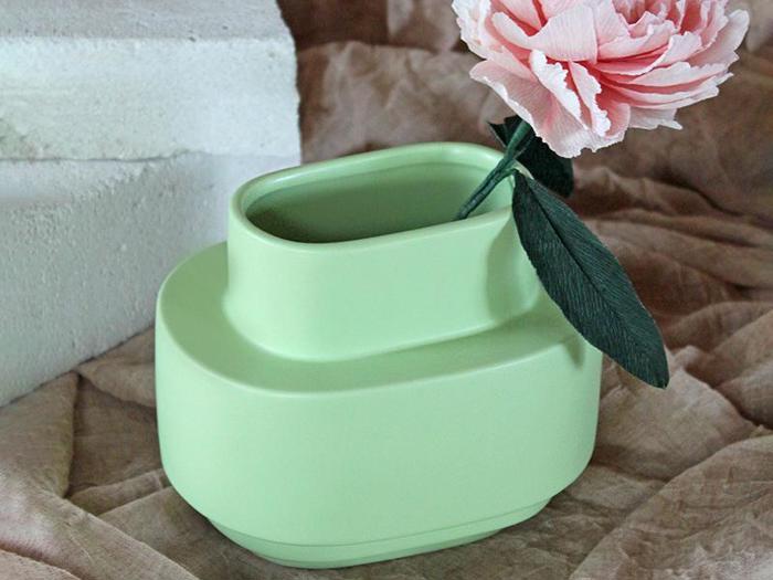 En pastelgrøn, moderne keramikvase rummer en enkelt lyserød kunstig blomst med en grøn stilk, der hviler på en beige stofbaggrund.