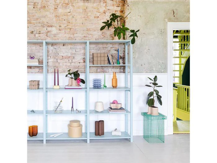 En lyseblå reol rummer dekorationsgenstande og planter mod en murstens- og stukvæg; en potteplante står på et grønt metalstativ nær en døråbning.