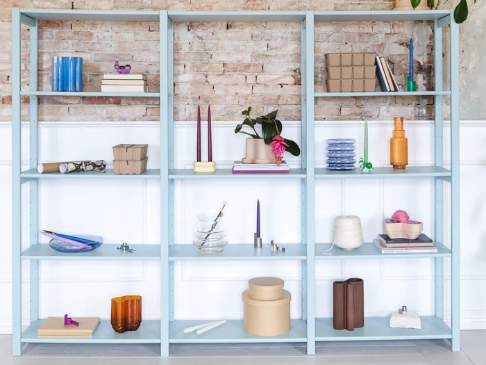 Lyseblå metalreol med pyntegenstande, bøger, stearinlys, glas og planter på en murstensvæg med hvid vægbeklædning.