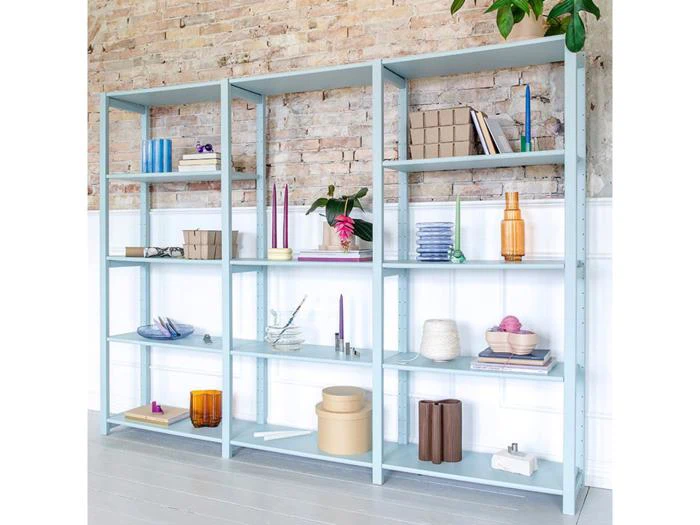 Lyseblå reol med forskellige pyntegenstande, bøger og vaser arrangeret pænt mod en murstensvæg og hvid vægbeklædning. En grøn plante hænger i øverste højre hjørne.