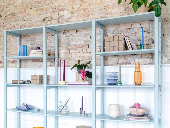Lyseblå metalhylder mod en murstensvæg rummer bøger, stearinlys, planter, keramik og dekorative genstande i pastelfarver, arrangeret pænt og sparsomt.