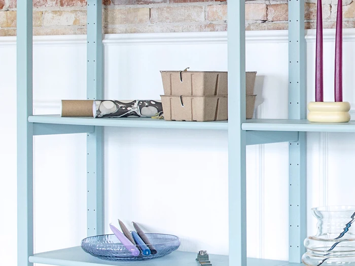 En lyseblå reol med madbeholdere af pap, en blå glasskål med lilla lys og høje lilla lys i en cremefarvet holder. Mursten og hvide vægge er synlige bagved.