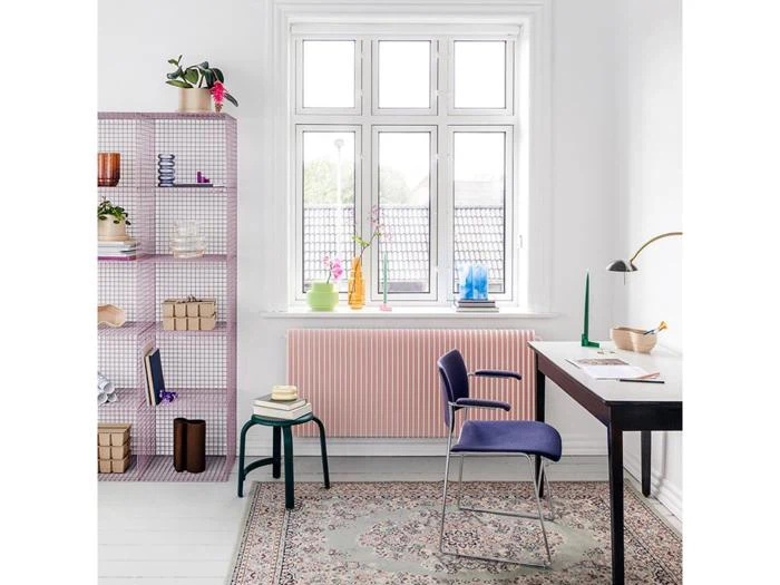 Et lyst, moderne arbejdsværelse med et stort vindue, et skrivebord med en lampe, en blå stol, en pink radiator, et mønstret tæppe og en lilla bogreol med farverige vaser og dekorationer.