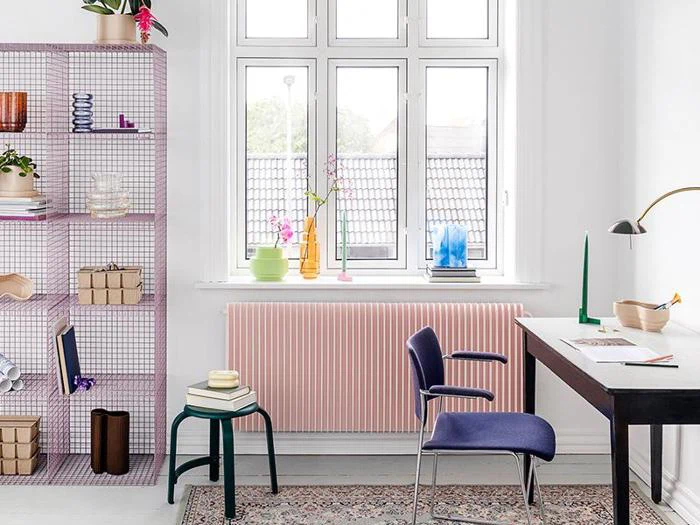 Et lyst, moderne hjemmekontor med sort skrivebord, blå stol, grøn skammel, pink trådhylder og farverig indretning foran et stort vindue med hvide vægge og et mønstret tæppe.