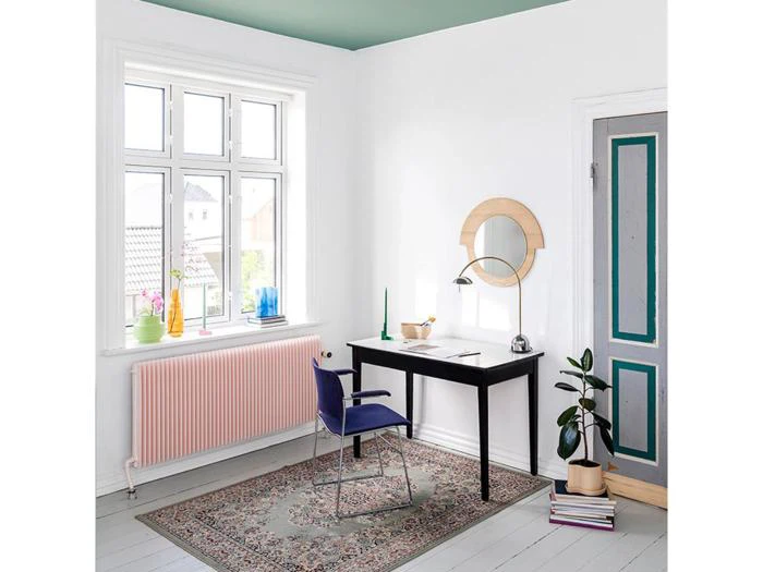 Minimalistisk hjemmekontor med sort skrivebord, blå stol, rundt spejl, potteplante og farverig dekoration i vindueskarmen. Stort vindue og blågrønt loft tilfører naturligt lys og farve.