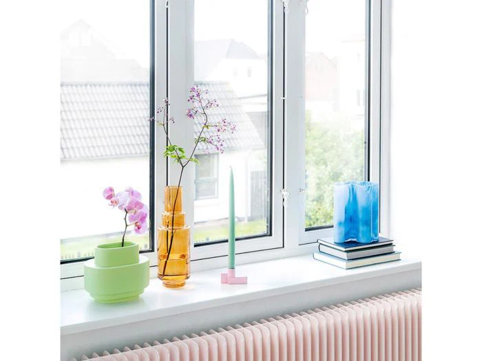 En lys vindueskarm med pastel- og glasvaser med blomster, en grøn rund potte, en blå vase på stablede bøger og et grønt lys med en lyserød holder over en lyserød radiator.