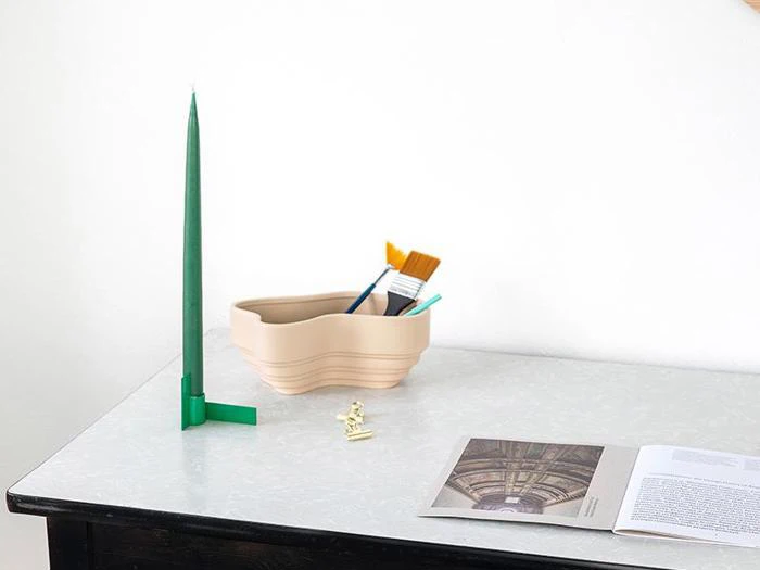 En hvid bordplade med et grønt, højt stearinlys, en beige beholder med pensler, en lille skakbrik og en åben bog med billeder og tekst.