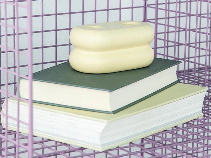To lukkede bøger stablet oven på hinanden i en lilla trådhylde med en beige, afrundet genstand placeret ovenpå.