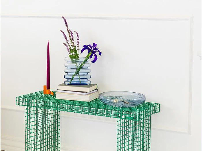 På et grønt ståltrådsbord står en glasvase med lilla blomster, en stak bøger, et rødt lys i en orange holder og en klar glasskål op ad en hvid væg.