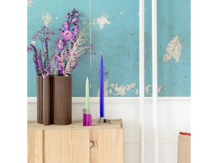 En moderne vase med lilla blomster og to høje stearinlys står på et træskab mod en blå, forvitret væg med afskallet maling og hvide rør.