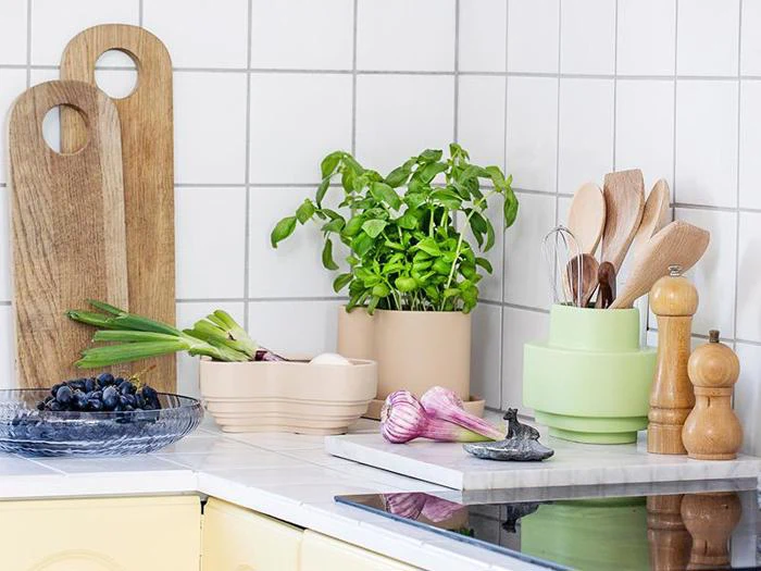 Et køkkenbord med skærebrætter, basilikum i potte, træredskaber, salt- og peberkværne, hvidløg, forårsløg, æg og en skål med vindruer mod en flisebelagt væg.